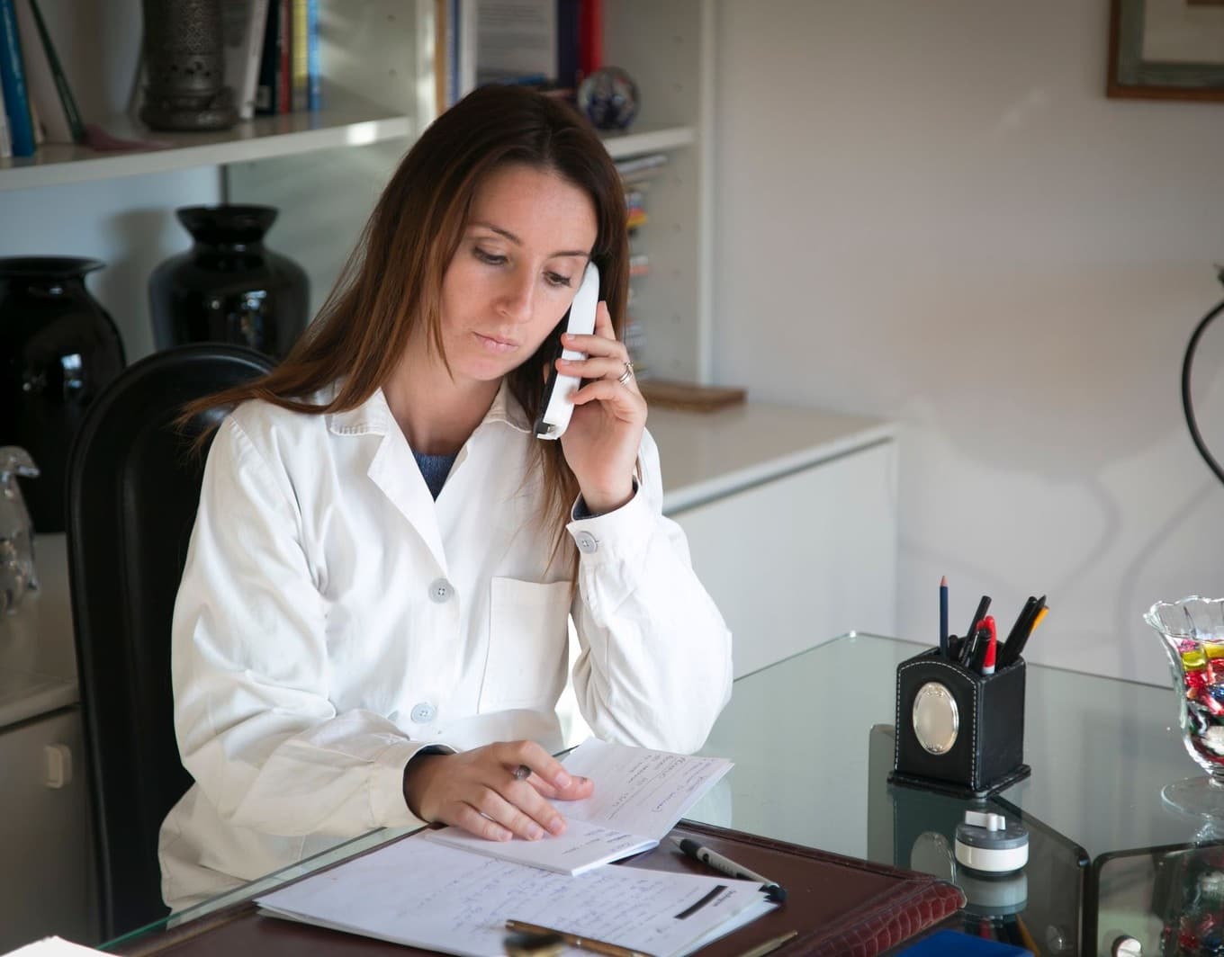 Doctor in a white coat on the phone at her desk, taking notes